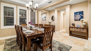 Un coin repas avec une table en bois dressée pour six personnes, flanquée de deux fenêtres équipées de stores, d’un lustre et d’une console sur laquelle sont posés des paniers.