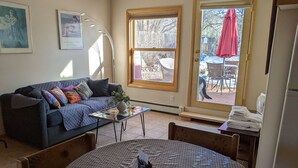 A living room with a blue sofa, a coffee table, and a dining table with chairs. There is a window with a view of the outside, a door, and a small table with towels. - Studio apartment 15 minute walk to the plaza (Santa Fe)