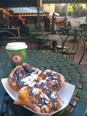 Una mesa con un pastel y una taza de café, rodeada de sillas y mesas de metal en un espacio exterior.