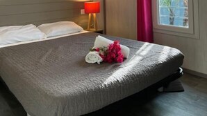 A hotel room with a bed, a nightstand, a window with a curtain, and a view of greenery outside.