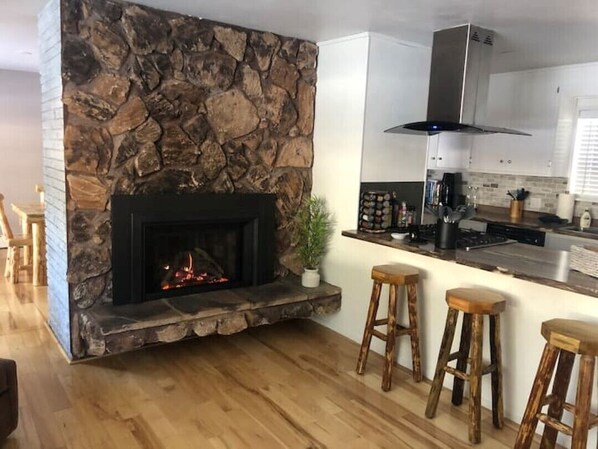 A modern kitchen with a stone fireplace, wooden bar stools, and a stainless steel range hood.