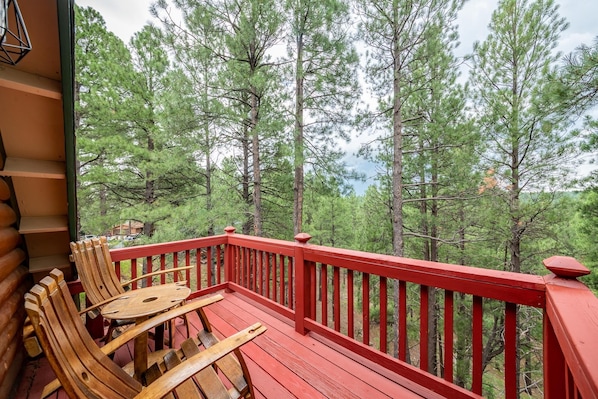A wooden deck with red railings and a table with chairs, overlooking a forest of tall pine trees.