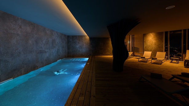 A modern indoor pool area with a wooden deck, lounge chairs, and a large tree trunk centerpiece. - Pena D'Água Boutique Hotel & Villas  (Covilhã)