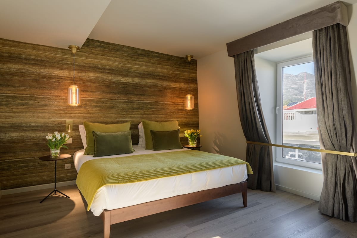 Ein modernes Schlafzimmer mit einer Wand in Holzoptik, einem großen Bett mit grüner Bettwäsche, zwei Pendelleuchten, einem Nachttisch mit Blumen und Blick auf ein Gebäude mit roten Dächern durch ein Fenster mit Vorhängen.