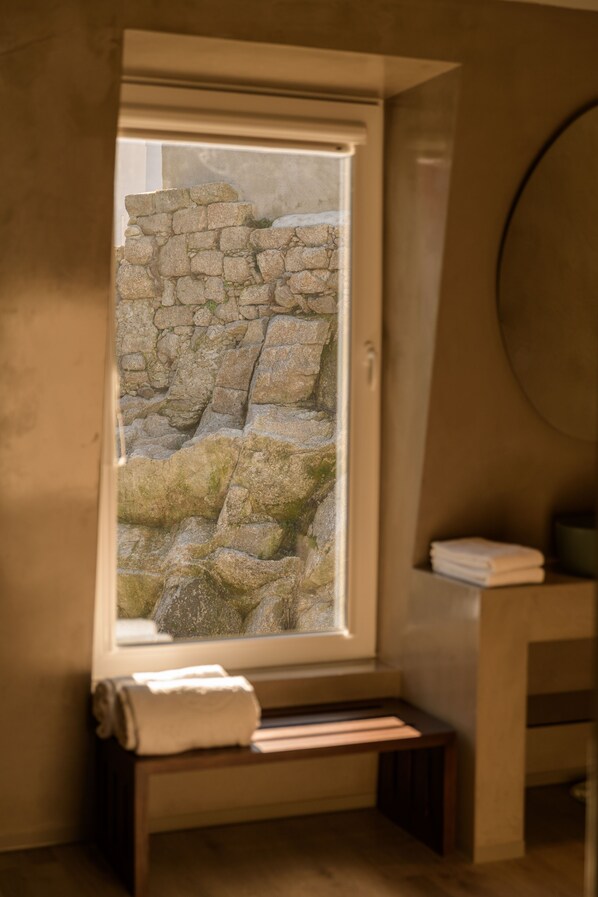A bathroom with a stone wall view, a bench with a towel, and a round mirror.