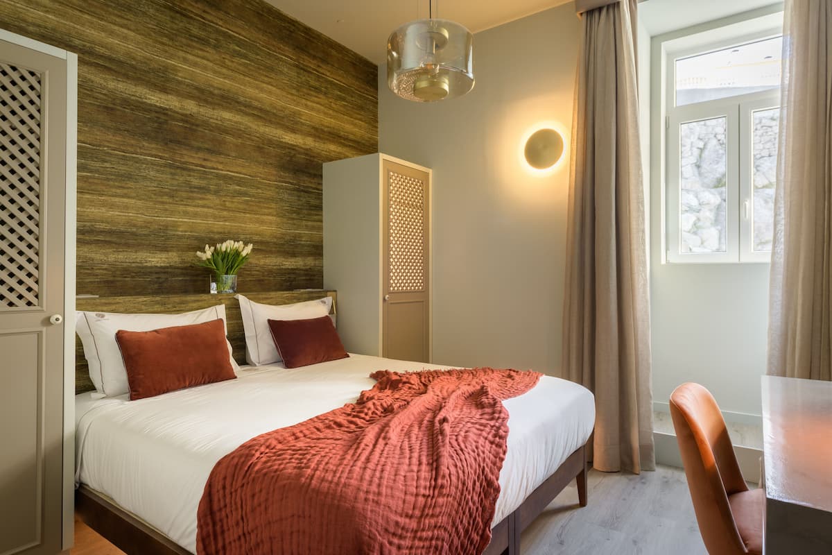 Ein ordentlich bezogenes Bett mit roter Decke, ein Holz-Kopfschild, ein Nachttisch mit Blumen in einer Vase und ein Fenster mit Vorhängen.