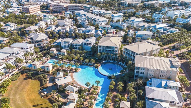 Eine Vogelperspektive zeigt einen Urlaubsort mit mehreren Gebäuden, einem zentral gelegenen Pool und üppigen Palmen.