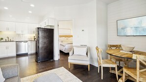 Eine moderne Küche mit weißen Schränken und einem schwarzen Kühlschrank, eine Essecke mit Holztisch und -stühlen sowie ein Schlafzimmer mit Bett und Nachttisch.