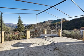 A covered outdoor area with a table and chairs, a metal railing, and a view of mountains.