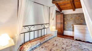 A bedroom with a canopy bed, a stone wall, a wooden ceiling, and a white dresser.