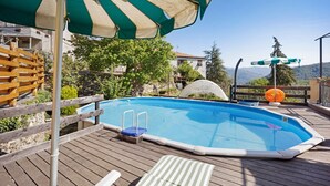 Una piscina exterior amb un tram inclinat de fusta, una tanca de fusta i un parasol verd i blanc a ratlles.