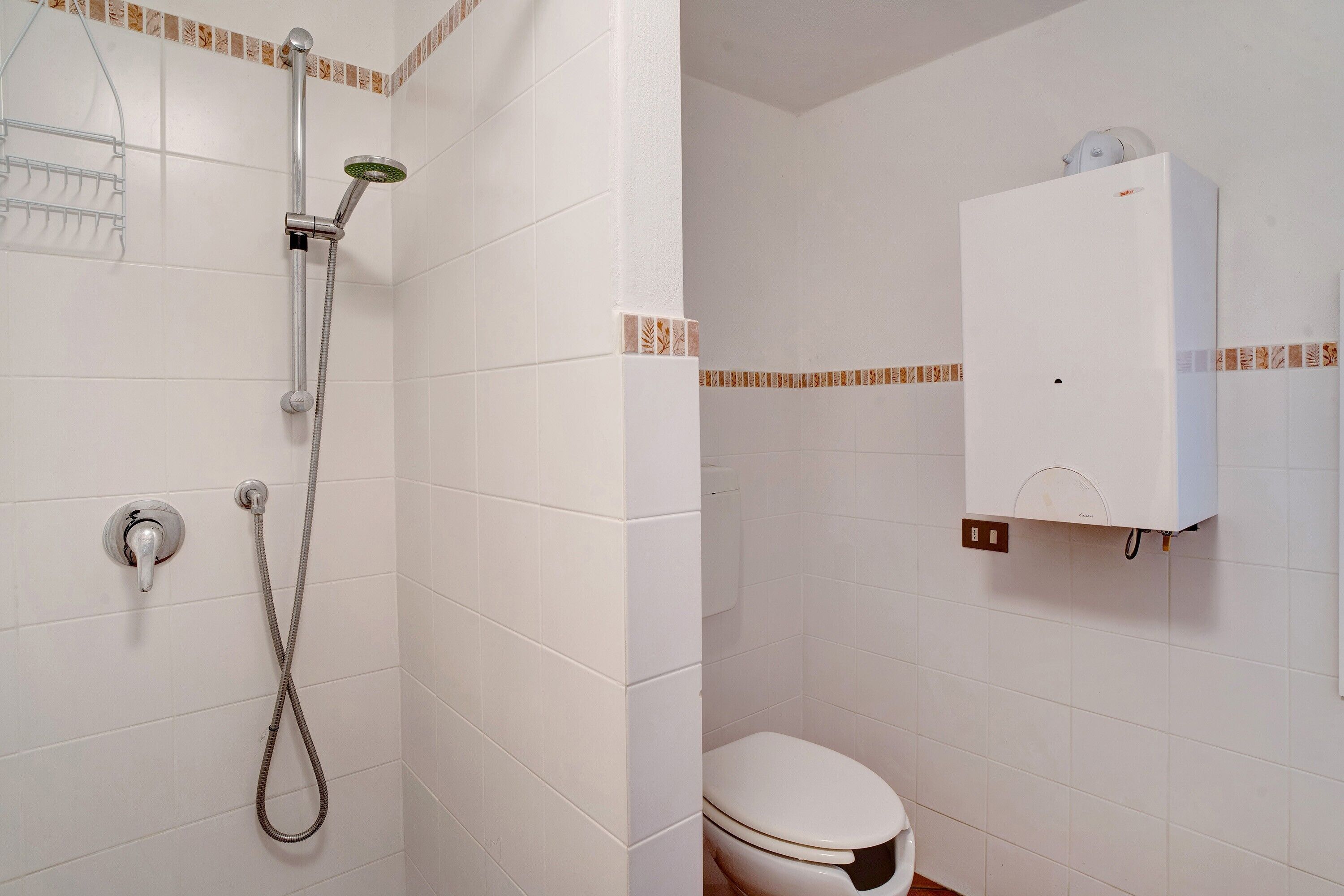 Une salle de bain avec des murs recouverts de carreaux blancs, une douche avec un pommeau flexible, des toilettes et un meuble blanc.