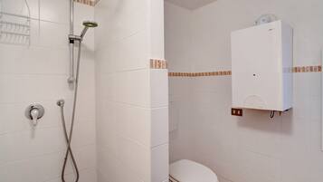 Une salle de bain avec des murs recouverts de carreaux blancs, une douche avec un pommeau flexible, des toilettes et un meuble blanc.