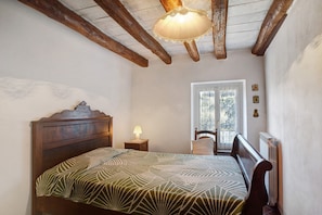 Un dormitorio con cabecera de madera, colcha estampada, mesita de noche con lámpara, radiador y ventana con vistas a árboles.