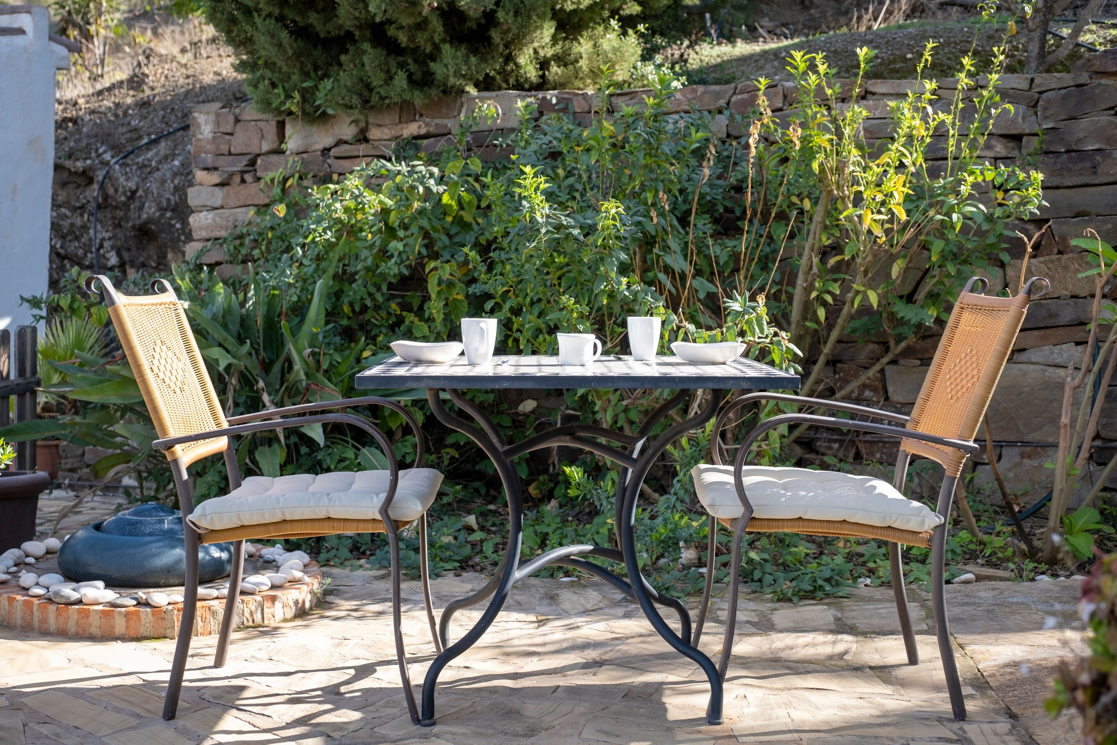 Un patio con una mesa redonda y dos sillas de mimbre, rodeado de follaje y un muro de piedra.