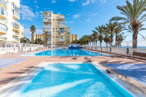 Una piscina bajo un cielo azul despejado, con palmeras y un edificio de varios pisos al fondo.