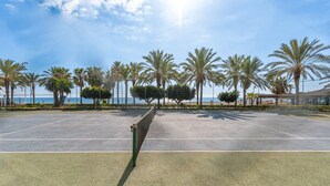 Un court de tennis en plein air, doté d’un filet, entouré de palmiers et surplombé d’un ciel dégagé.