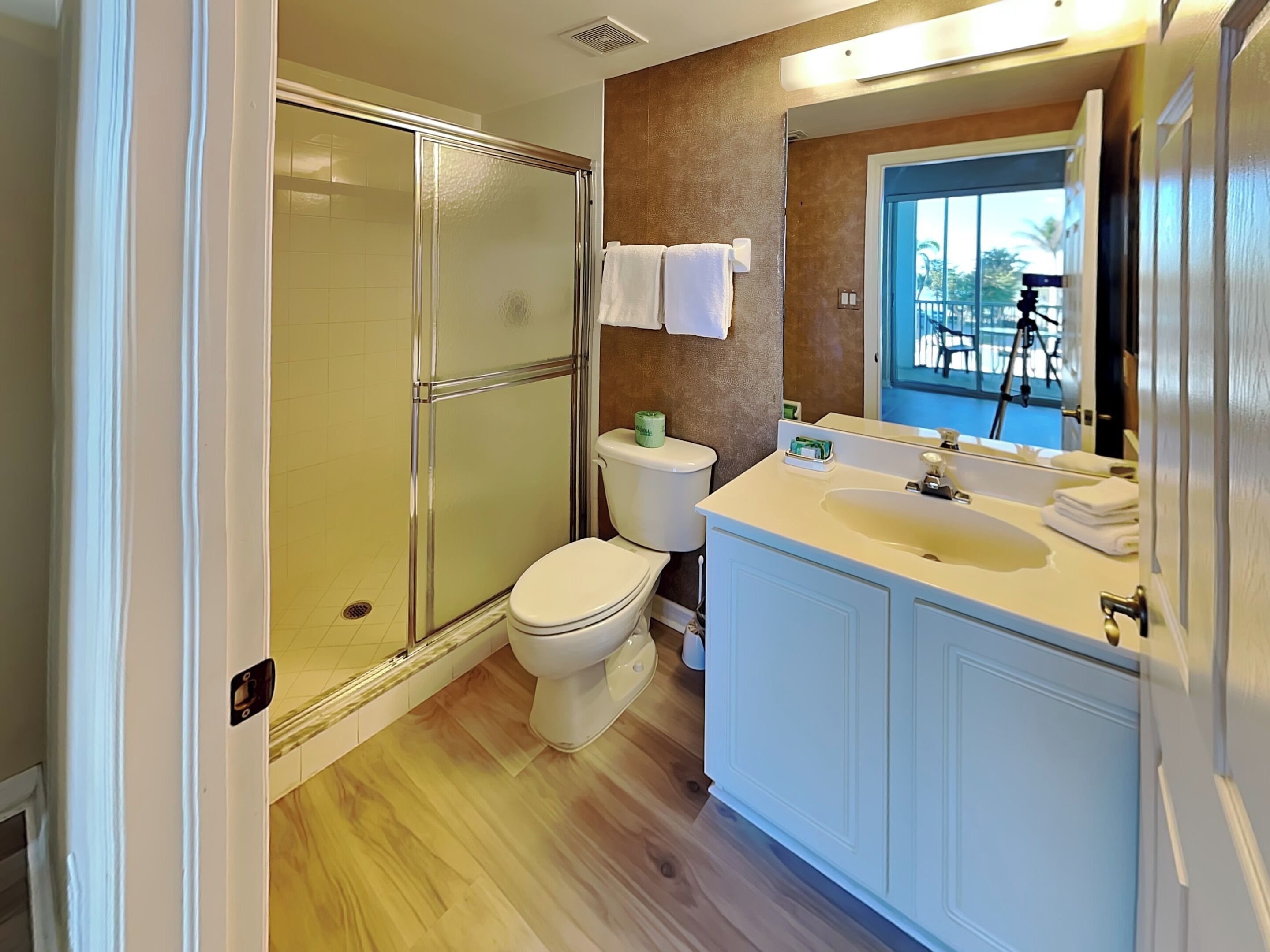 Un baño con cabina de ducha de vidrio, inodoro, lavabo con espejo y ventana con vistas a palmeras.