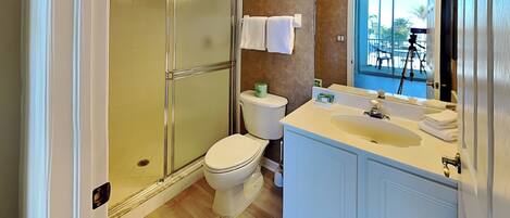 Un baño con cabina de ducha de vidrio, inodoro, lavabo con espejo y ventana con vistas a palmeras.