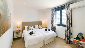 Een slaapkamer met twee bedden, een nachtkastje met een lamp, een stoel en een groot raam met gordijnen.