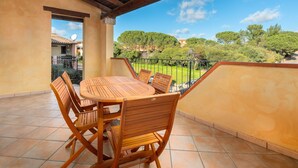Une terrasse couverte avec une table et des chaises en bois, d’où l’on peut admirer des arbres et des bâtiments en arrière-plan.