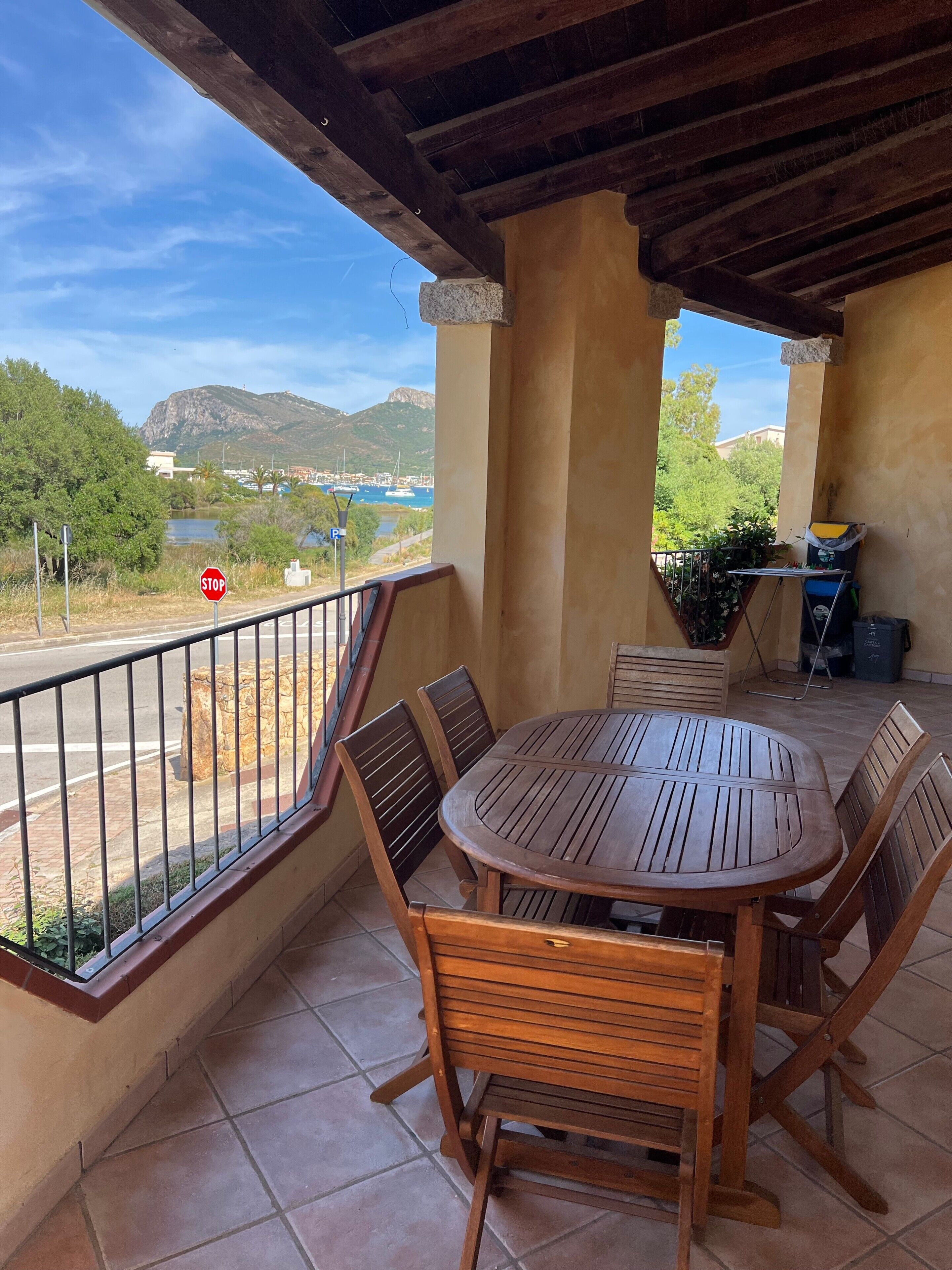 Un patio al aire libre cubierto con una mesa y sillas de madera redonda, vista a un cuerpo de agua y colinas, y una señal de stop al fondo.