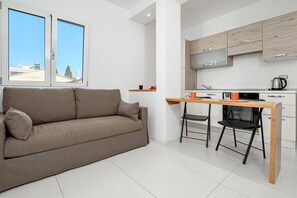 A modern apartment with a sofa, a dining table, chairs, a kitchen area, and a view through large windows.