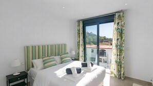 A bedroom with a large bed, striped headboard, pillows, a nightstand, a lamp, and a view of a building through a window with curtains. - Stunning Apartment in Cómpeta (Cómpeta)