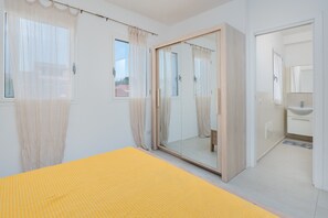 Un dormitorio con cama, una puerta corredera, un baño con lavamanos y una ventana con cortinas translúcidas.