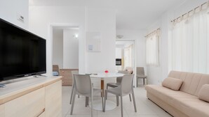 Una sala de estar moderna con un televisor de pantalla plana, una mesa de comedor redonda con sillas, un sofá color beige y una cocina al fondo.