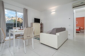 Un salon moderne avec un canapé blanc, une table à manger avec des chaises, un téléviseur posé sur un meuble et une vue sur l’extérieur.