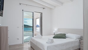 A bedroom with a bed, a nightstand, a lamp, a green pillow, a window with a view of the sea, and a sliding glass door. - Villa "Cormorani 1" with Sea View, Jacuzzi & Wi-Fi (golfo aranci)