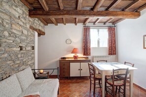 Une pièce rustique avec des murs en pierre, des poutres en bois, une table à manger avec des chaises, un canapé et une fenêtre avec des rideaux.