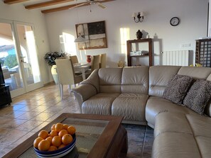 Ein Wohnzimmer mit einem beigefarbenen Sofa, einem Couchtisch mit Orangen, einem Essbereich mit Stühlen, einem Deckenventilator und einer Uhr an der Wand.