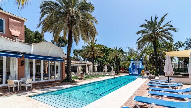 Piscina con sillones de descanso, palmeras y un edificio con toldo azul.
