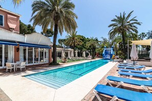 A swimming pool with lounge chairs, palm trees, and a building with a blue awning. - Beautiful Apartment in Miraflores with Sea View, Shared Pool and Wi-Fi (Mijas Costa)