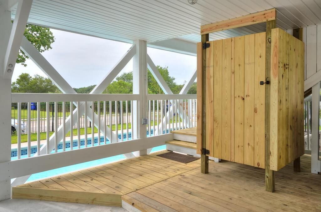 Un espacio al aire libre techado con piso de madera, barandilla blanca y vista a una piscina.
