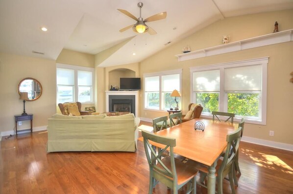 A spacious living room with a fireplace, a dining area with wooden table and chairs, and large windows with blinds.