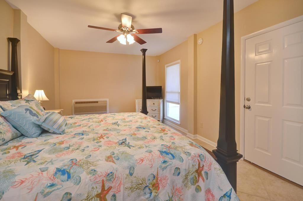 Quarto com cama, ventoinha no teto, mesa de cabeceira, cômoda e porta.