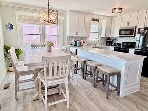 A modern kitchen with white cabinetry, a central island with bar stools, a chandelier, and a dining area with chairs. - Oceanfront retreat, Sea-ing Starrs in Oak Island, offers 5 bedrooms for 10 guests. (Oak Island)