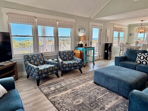 A living room with a TV, two patterned armchairs, a sofa, a rug, and a kitchen area with a fridge and a table. - Oceanfront retreat, Sea-ing Starrs in Oak Island, offers 5 bedrooms for 10 guests. (Oak Island)