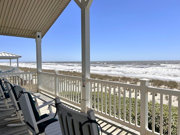 A beach view from a covered deck with white railings and several rocking chairs.