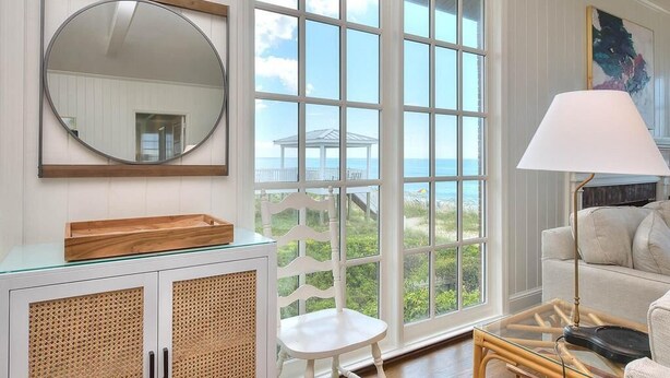 Une pièce avec une grande fenêtre, un miroir rond, une chaise blanche, une table avec un plateau en verre et une lampe.
