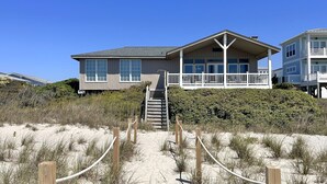 Une maison située en bord de mer, dotée d’un balcon, d’un escalier menant à l’entrée et d’une plage de sable en premier plan.