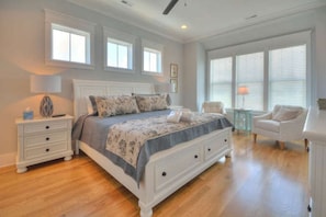 A bedroom with a large bed, a nightstand, a chair, and a window with blinds. - A Perfect Getaway: Private Pool, Elevator, Walk to Beach (Oak Island)
