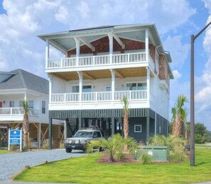 A two-story building with a balcony on the upper floor, a vehicle parked in front, and a sign indicating a nearby location. - A Perfect Getaway: Private Pool, Elevator, Walk to Beach (Oak Island)