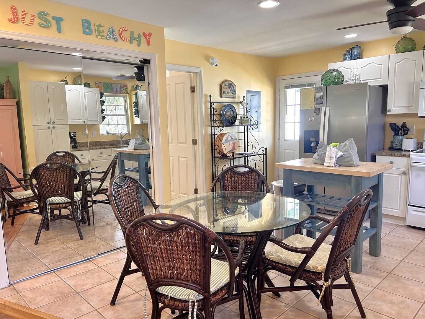Un coin repas avec des chaises en osier et une table en verre, une cuisine avec des meubles blancs et un panneau sur lequel on peut lire : « JUST BEACHY ».