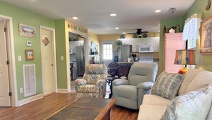 A living room with a wooden coffee table, a beige sofa, and patterned armchairs. A kitchen is visible in the background with white cabinets and a microwave.