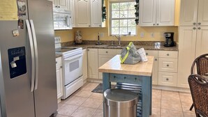 A kitchen with white cabinets, a stainless steel refrigerator, a microwave, a stove, and a sink. There is a window with a view of trees, a ceiling fan, and a trash can.
