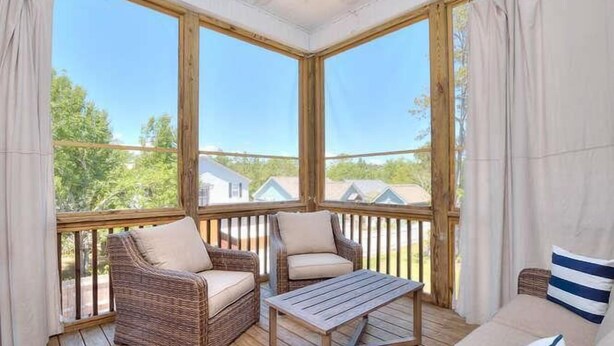 Une véranda meublée de mobilier en osier, avec une table en bois et de grandes fenêtres donnant sur des maisons et la verdure.